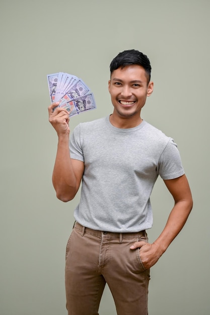 Hombre asiático sonriente sosteniendo billetes de dólar en efectivo mientras está de pie contra un fondo de estudio verde