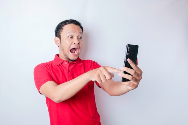 Hombre asiático sorprendido con camiseta roja apuntando a su teléfono inteligente aislado de fondo blanco