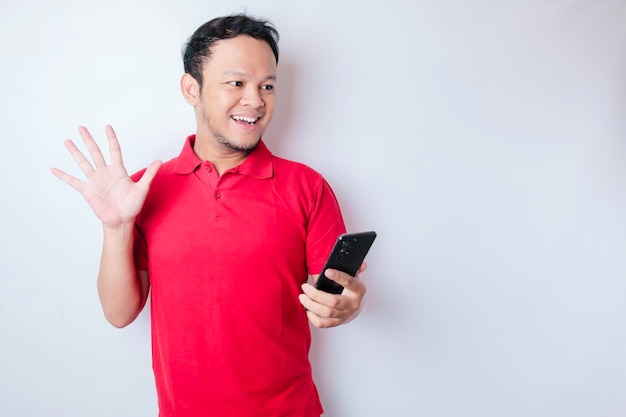 Hombre asiático sorprendido con camiseta roja apuntando a su teléfono inteligente aislado de fondo blanco