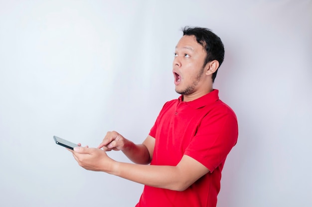 Hombre asiático sorprendido con camiseta roja apuntando a su teléfono inteligente aislado de fondo blanco