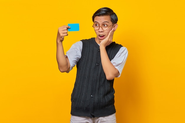 Hombre asiático sorprendido que sostiene la tarjeta de crédito con la boca abierta y la mano en una mejilla sobre fondo amarillo
