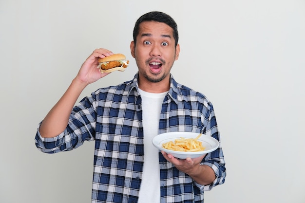 Hombre asiático sosteniendo una hamburguesa y papas fritas con expresión asombrada