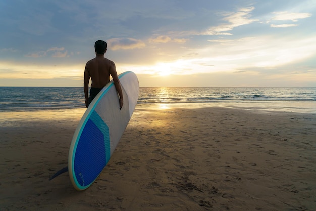 Hombre asiático sosteniendo una tabla de sup y remando y caminando en la playa En el fondo el océano y la puesta de sol Vista posterior Surf de verano