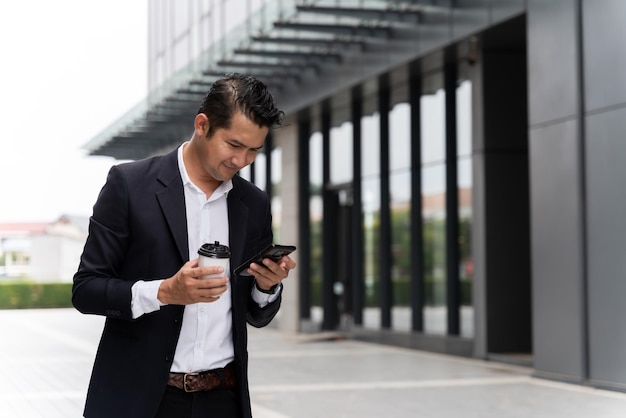 Foto hombre asiático sosteniendo una taza de café y usando un teléfono profesionales interactuando con sus dispositivos en espacios públicos urbanos