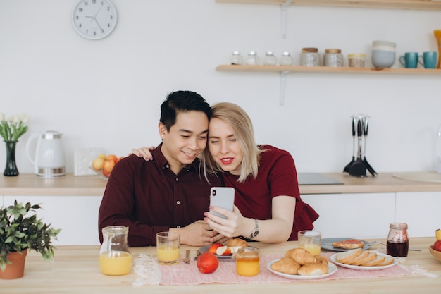 El hombre asiático y su mujer rubia caucásica desayunan. Pasan tiempo juntos en la cocina y miran videos en el teléfono.