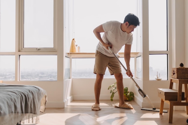 Foto hombre asiático tailandés usando una mopa para limpiar el suelo en el apartamento de la sala de estar hombre haciendo tareas domésticas