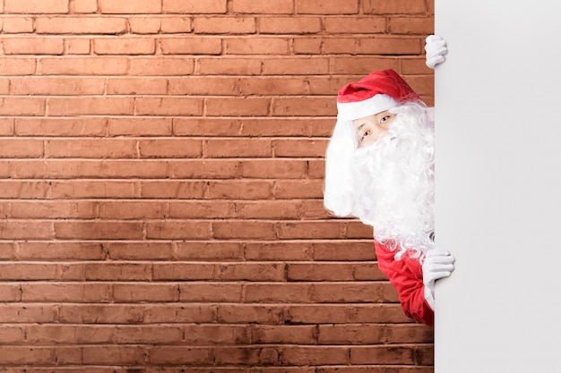 Hombre asiático en traje de Santa mostrando un tablero vacío con una pared de ladrillos.