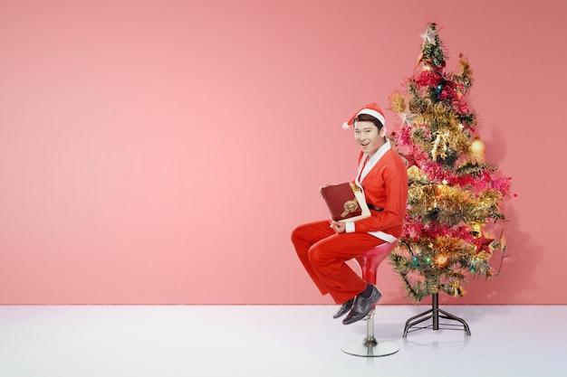 Hombre asiático en traje de Santa sentado y sosteniendo la caja de regalo con árbol de Navidad decorado con luces de colores y adornos.