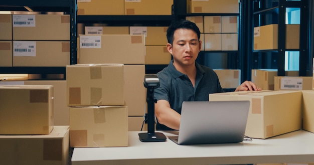 Foto el hombre asiático usa datos de escaneo de código de barras en la caja y escribe el pedido en línea de registro en la computadora portátil en el almacén