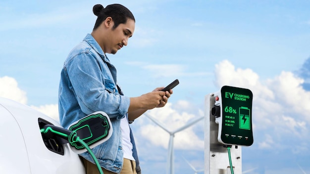 Hombre asiático usando un teléfono inteligente mientras un automóvil eléctrico recarga energía desde un cargador EV desde una estación de carga en un parque de turbinas eólicas Concepto alternativo de utilización de energía limpia y sostenible