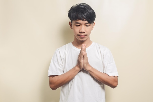 Hombre asiático vestido con camiseta blanca con gesto de oración sobre fondo aislado