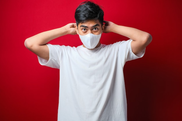 Hombre asiático vestido con camiseta blanca poniéndose una mascarilla con una mano poniendo la banda elástica en una oreja sobre fondo rojo.