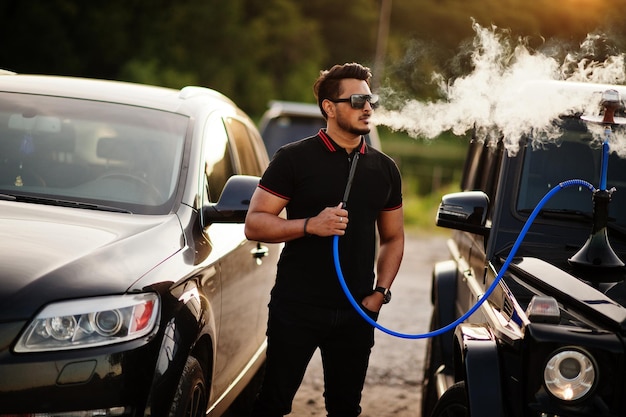 El hombre asiático viste todo negro posado cerca de un auto todoterreno y fuma narguile.