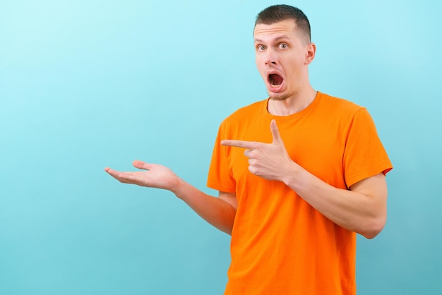 Un hombre asombrado con la boca abierta en una camiseta naranja apuntando con una mano y señalando con el dedo en el lado medio Mostrando espacio de copia vacío sobre fondo azul Guapo modelo alegre presente