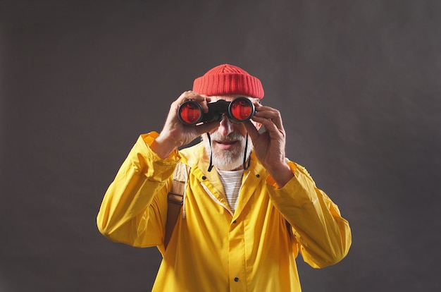 Un hombre asombroso, un hombre con una gorra roja y un impermeable amarillo brillante disfruta de las vistas de smortya en la distancia usando binoculares, fondo aislado. Hobby