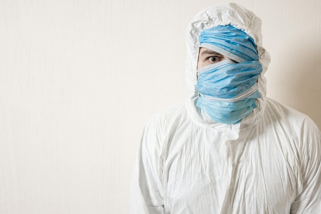 Un hombre asustado con un traje protector colgado con máscaras médicas representa el horror contra una pared blanca. Los horrores de la epidemia, el peligro del coronavirus