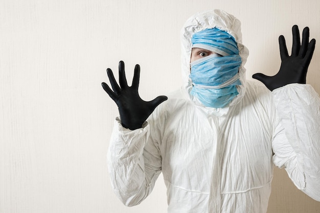 Un hombre asustado con un traje protector colgado con máscaras médicas representa el horror contra una pared blanca. Los horrores de la epidemia, el peligro del coronavirus