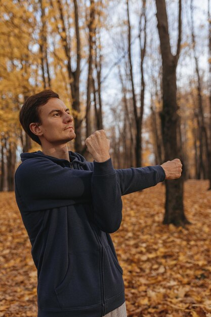 Hombre atlético estirando los brazos y calentando el cuerpo mientras hace ejercicio físico en el parque de otoño