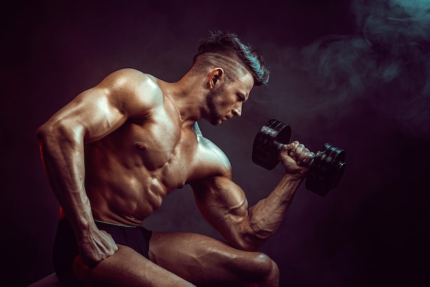 Hombre atlético flexionando los músculos en estudio sobre pared oscura con humo. Culturista fuerte con abdominales perfectos.
