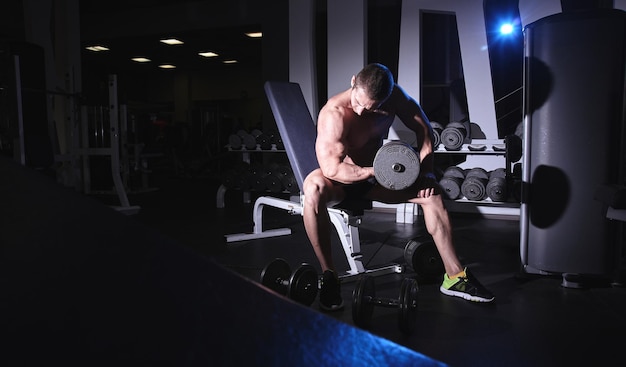 Hombre atlético fuerte musculoso con torso desnudo bombeando músculos de las manos con pesas en el gimnasio, entrenamiento brutal de culturista, nutrición deportiva y concepto de equipo