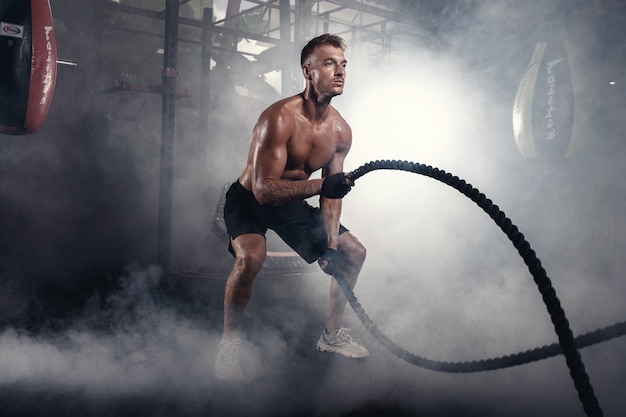 Hombre atlético haciendo ejercicios de crossfit con cuerda en el gimnasio ahumado