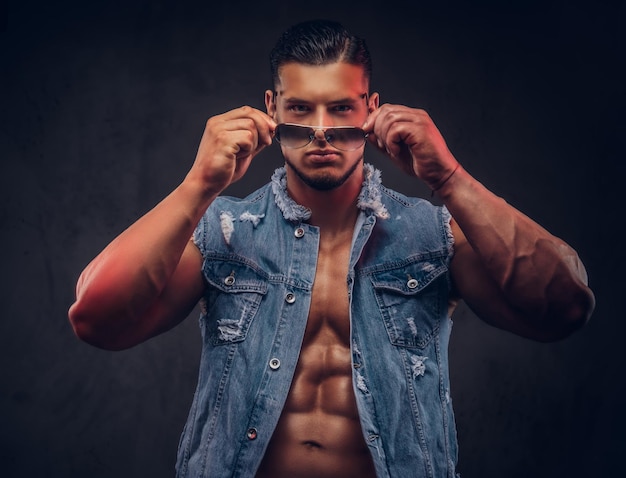 Hombre atlético de moda desnudo sexy en un chaleco de mezclilla y poses de gafas de sol en un estudio. Aislado sobre fondo oscuro.