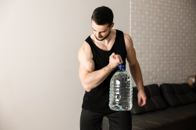 Hombre atlético que hace ejercicios de levantamiento de pantorrillas con una botella grande de agua. Habitación moderna blanca en el fondo. Hombre fuerte en ropa deportiva negra haciendo ejercicio para tener un cuerpo en forma.