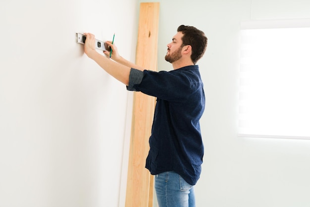 Hombre atractivo de unos 30 años usando un nivel para comprobar una nueva pared. Handyman trabajando en su casa para hacer algunas mejoras