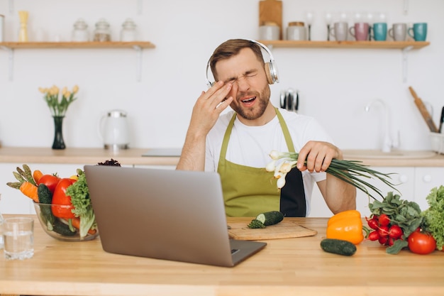 Hombre atractivo con auriculares sentado en la mesa en la cocina preparando ensalada con cebolla y limpiando los ojos de las lágrimas