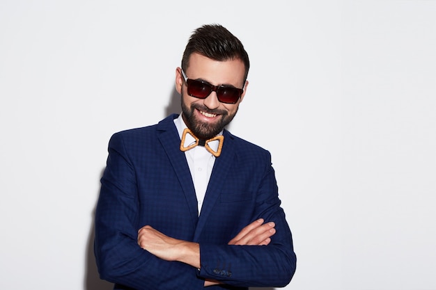 Hombre atractivo con cabello negro y barba con camisa blanca con pajarita, traje azul y gafas de sol en el fondo blanco del estudio, retrato, posando.