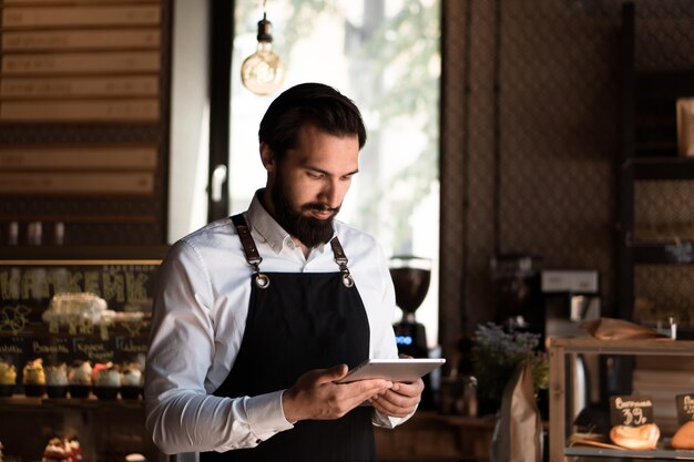 Foto hombre atractivo en delantal barista o propietario se encuentra en la cafetería con tableta