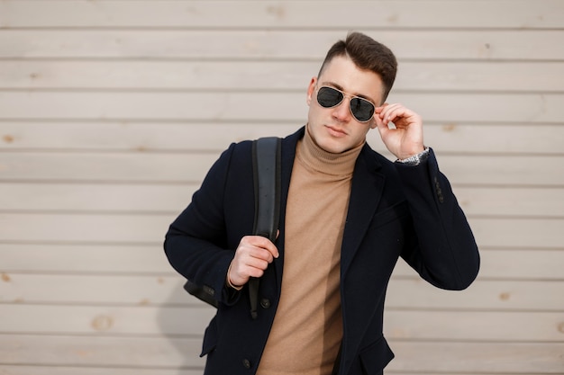 Hombre atractivo joven inconformista con gafas elegantes en una chaqueta de moda con un suéter de punto vintage con un peinado elegante con una mochila