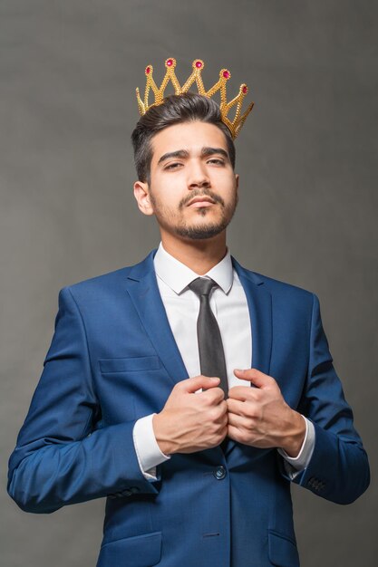 Foto hombre atractivo joven en un traje azul con una corona en la cabeza sobre un fondo gris