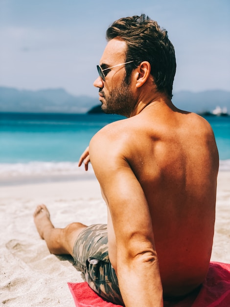 Hombre atractivo mirando a otro lado mientras está sentado en la playa de agua azul