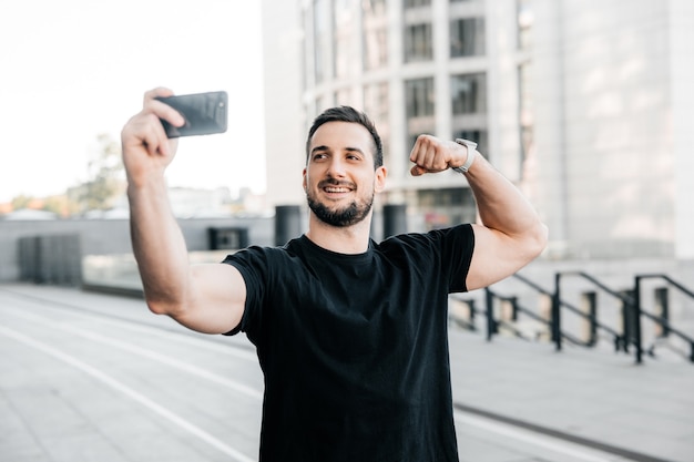 Hombre atractivo muestra bíceps y toma un selfie. Concepto de redes sociales. Atleta masculino en camiseta negra muestra a sus amigos en una videollamada su cuerpo en forma. Entrenamiento de la mañana. Concepto de deporte urbano.