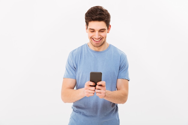 Hombre atractivo con el pelo oscuro corto chateando o escribiendo mensajes de texto con teléfono celular, aislado sobre la pared blanca