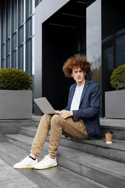 Hombre atractivo pensativo que usa una computadora portátil al aire libre Programador exitoso sentado cerca de la oficina moderna