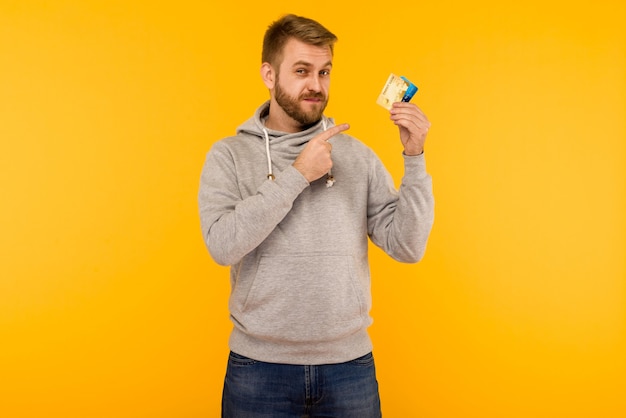 Hombre atractivo con una sudadera con capucha gris señala con el dedo la tarjeta de crédito que tiene en la mano sobre un fondo amarillo - imagen
