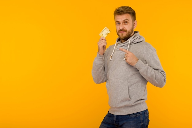 Hombre atractivo con una sudadera con capucha gris señala con el dedo la tarjeta de crédito que tiene en la mano sobre un fondo amarillo - imagen