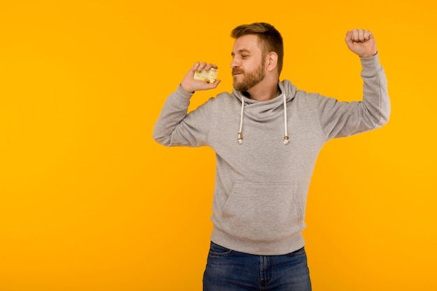 Hombre atractivo con una sudadera gris baila alegremente con una tarjeta de crédito en la mano sobre un fondo amarillo - imagen