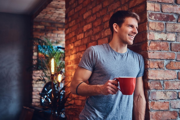 Un hombre atractivo tatuado con estilo vestido con jeans y una camiseta, se inclina contra la pared de ladrillo, sosteniendo una taza de café. Sonriendo y mirando a otro lado.