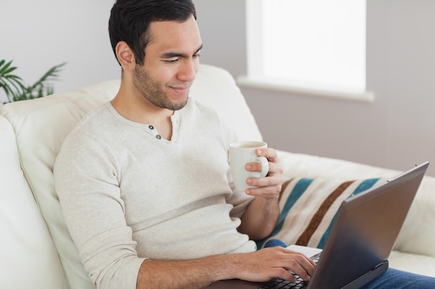 Hombre atractivo tranquilo tomando café mientras trabaja en su computadora portátil