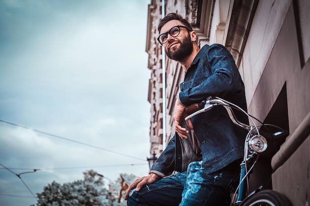 Un hombre atractivo con vaqueros y gafas está parado en la calle. Él está apoyado en su bicicleta. El hombre está mirando hacia arriba. Edificio, cielo y árbol al fondo.