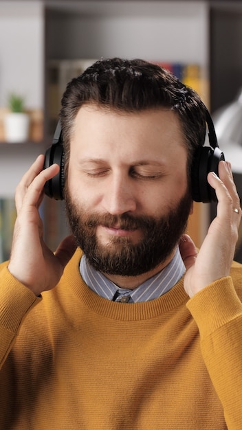 Hombre con auriculares escucha música. Vista vertical del hombre barbudo positivo en auriculares inalámbricos negros sostiene la cabeza con las manos y disfruta de la música. Tiro medio