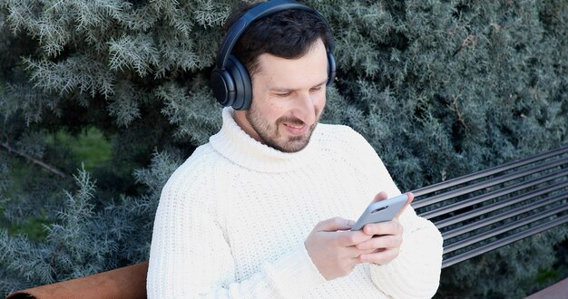 Un hombre con auriculares y un suéter blanco está sentado en un banco y mira una tableta.