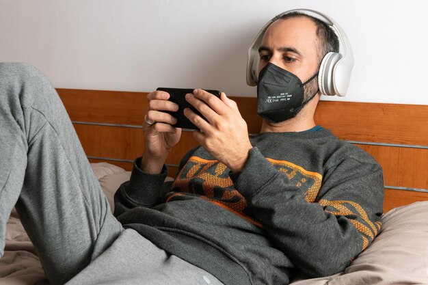 Hombre con auriculares, teléfono móvil y mascarilla, sentado en su cama y confinado en su habitación en casa