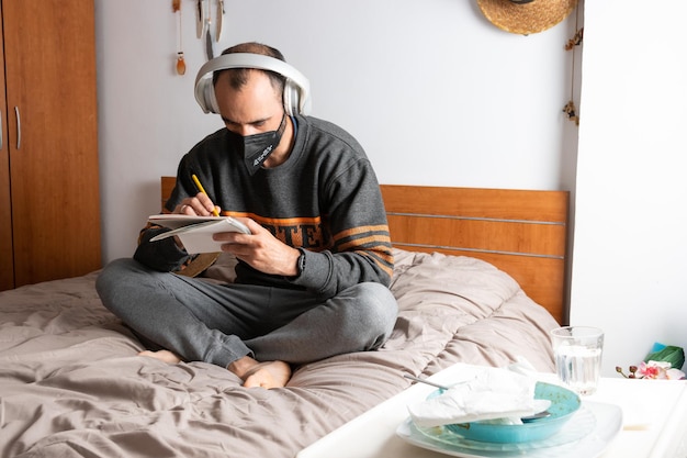 Hombre con auriculares, teléfono móvil y mascarilla, sentado en su cama y confinado en su habitación en casa