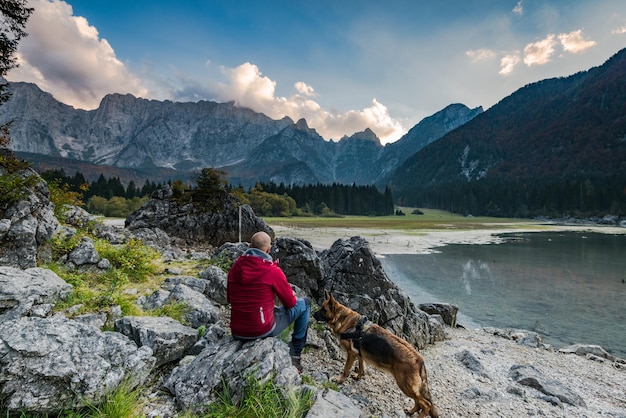 Hombre de aventuras con perro sentado en una roca mirando el lago alpino