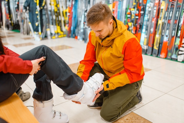 El hombre ayuda a la mujer a probarse botas de esquí o snowboard, ir de compras en la tienda de deportes. Estilo de vida extremo de la temporada de invierno, tienda de ocio activo, compradores que eligen proteger el equipo