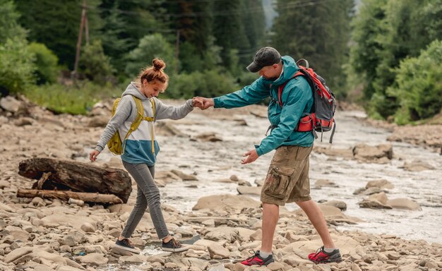 Hombre ayudando a la mujer joven a cruzar el río de las montañas rocosas durante el senderismo juntos